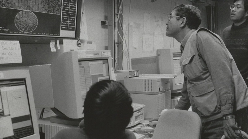 白黒の写真。戸塚さんがパソコンの前に立ってモニターを見上げている。